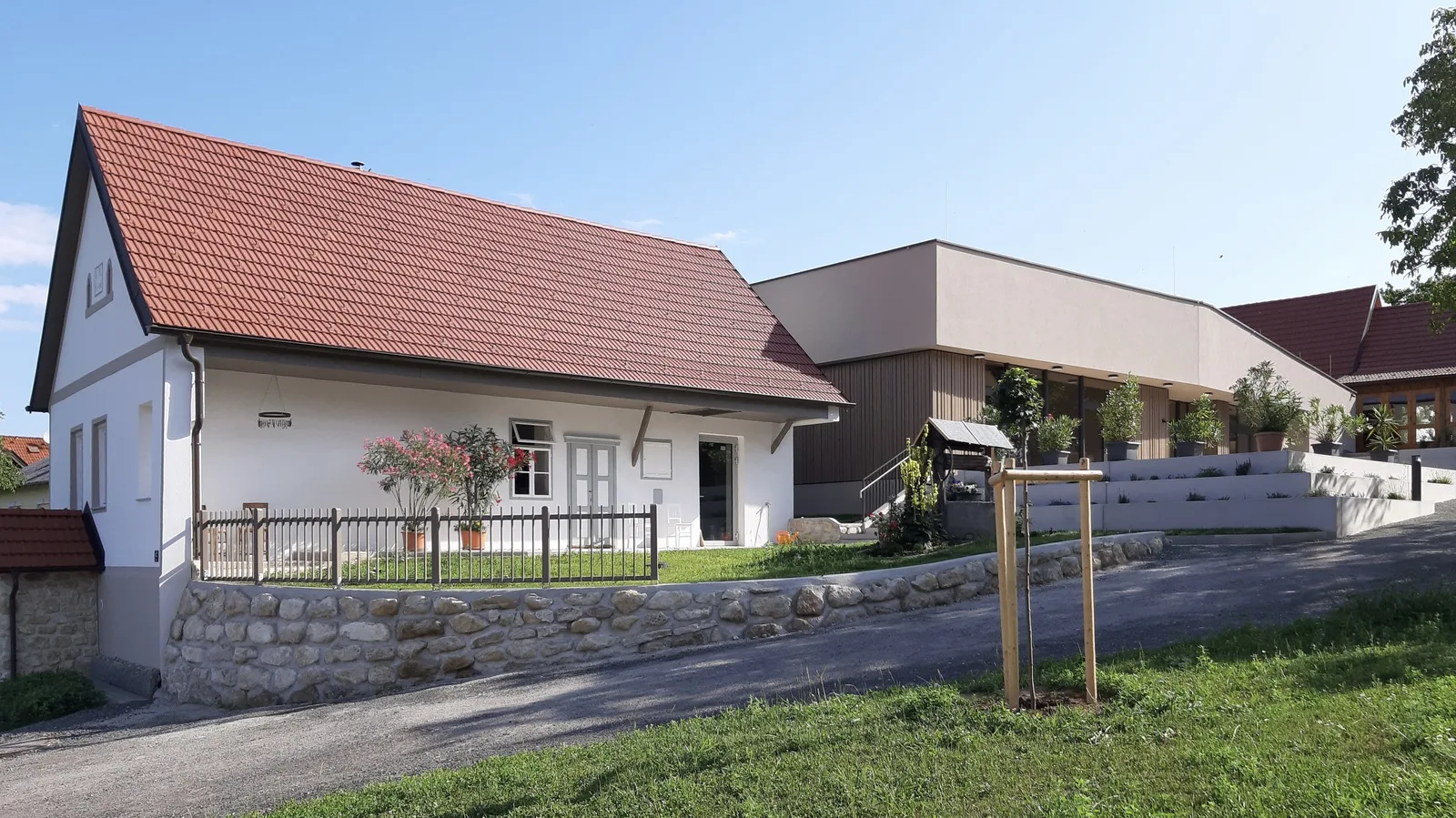 Bild eines alten weißen Bauernhauses mit Giebeldach, daneben ein zeitgenössischer Zubau in brauner Farbe