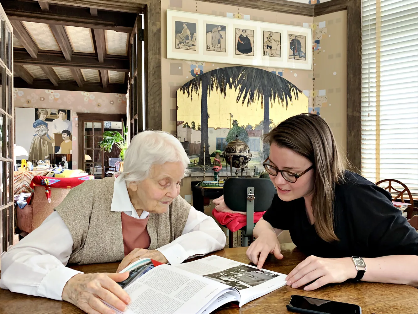 Die Architektin Denise Scott Brown sitzt gemeinsam mit der Autorin Frida Grahn blättern, an einem Tisch sitzend, in einem Buch.