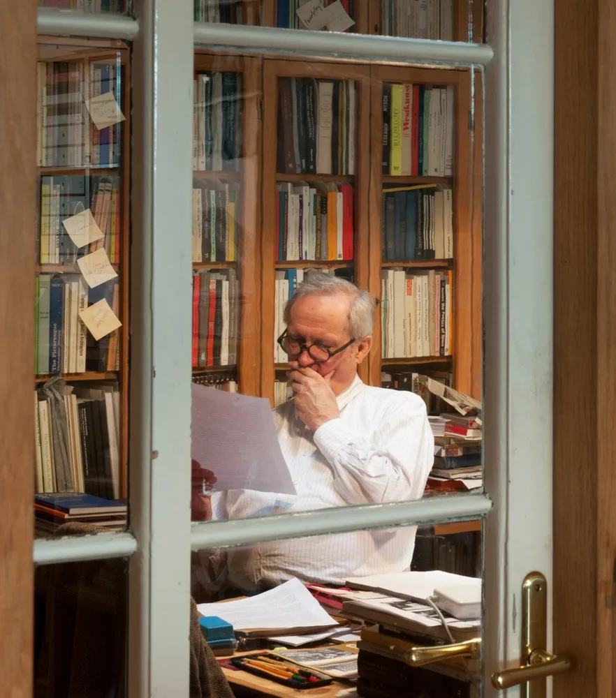 Porträt eines Mannes an seinem Schreibtisch, umgeben von Büchern und Bücherregalen, fotografiert durch ein Fenster