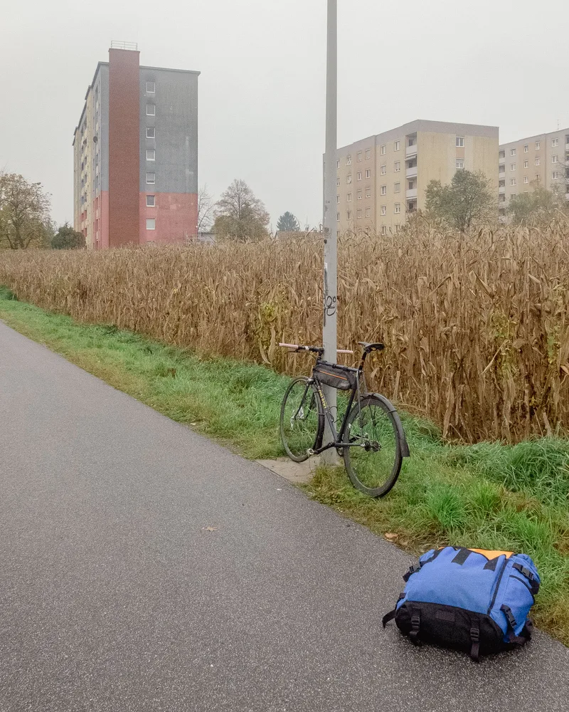 Foto eines Fahrrads am Straßenrand, vor einem Maisfeld. In Hintergrund drei Wohntürme im Nebel