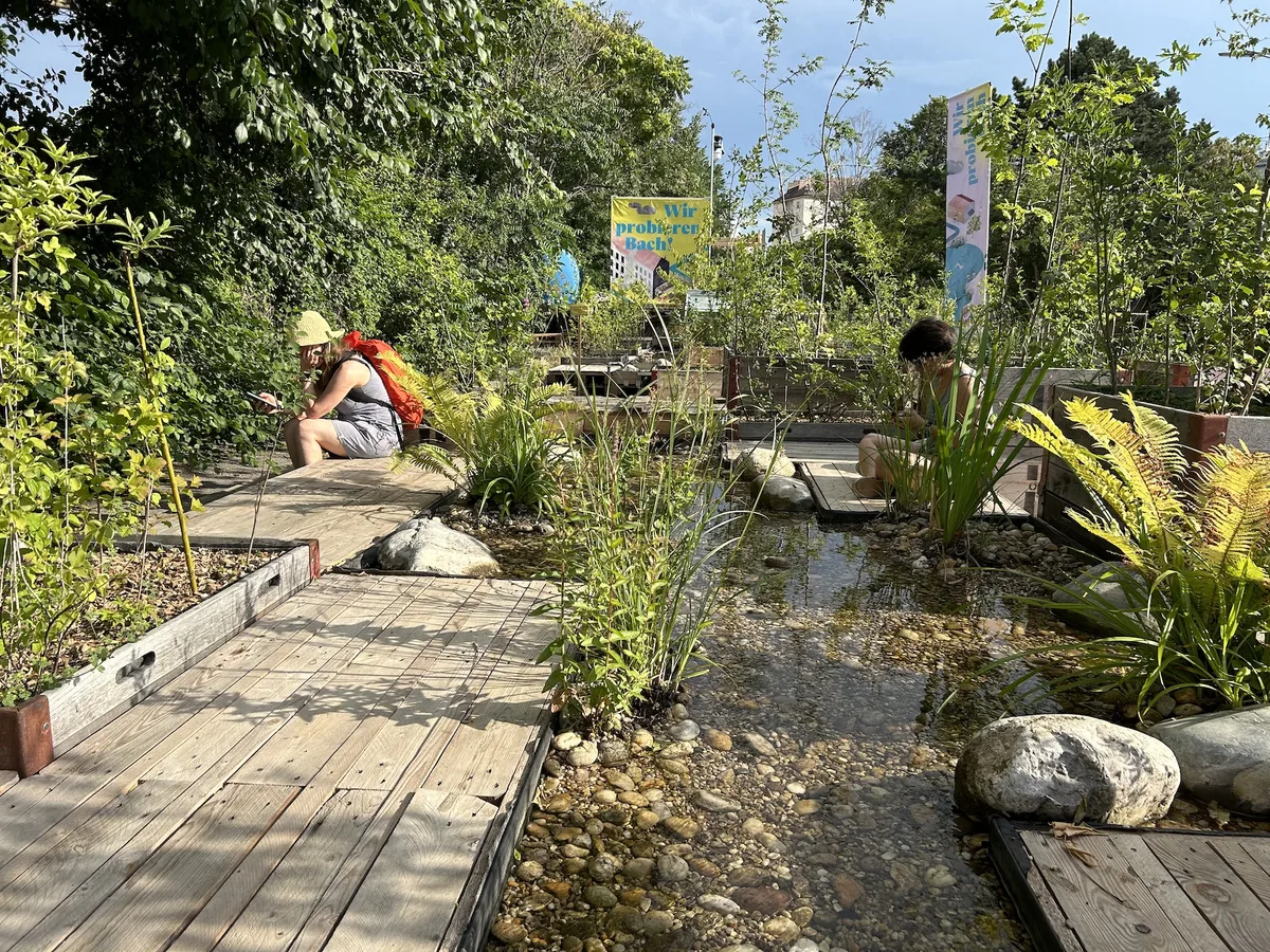 © Christine Müller
Quelle: Christine Müller Man sieht einen seichten Wasserlauf mit Kieselsteinen eingefasst mit Holzflächen und dicht bewachsenem Ufer bei strahlend blauem Himmel uns Sonnenschein, an dem zwei Personen sitzen