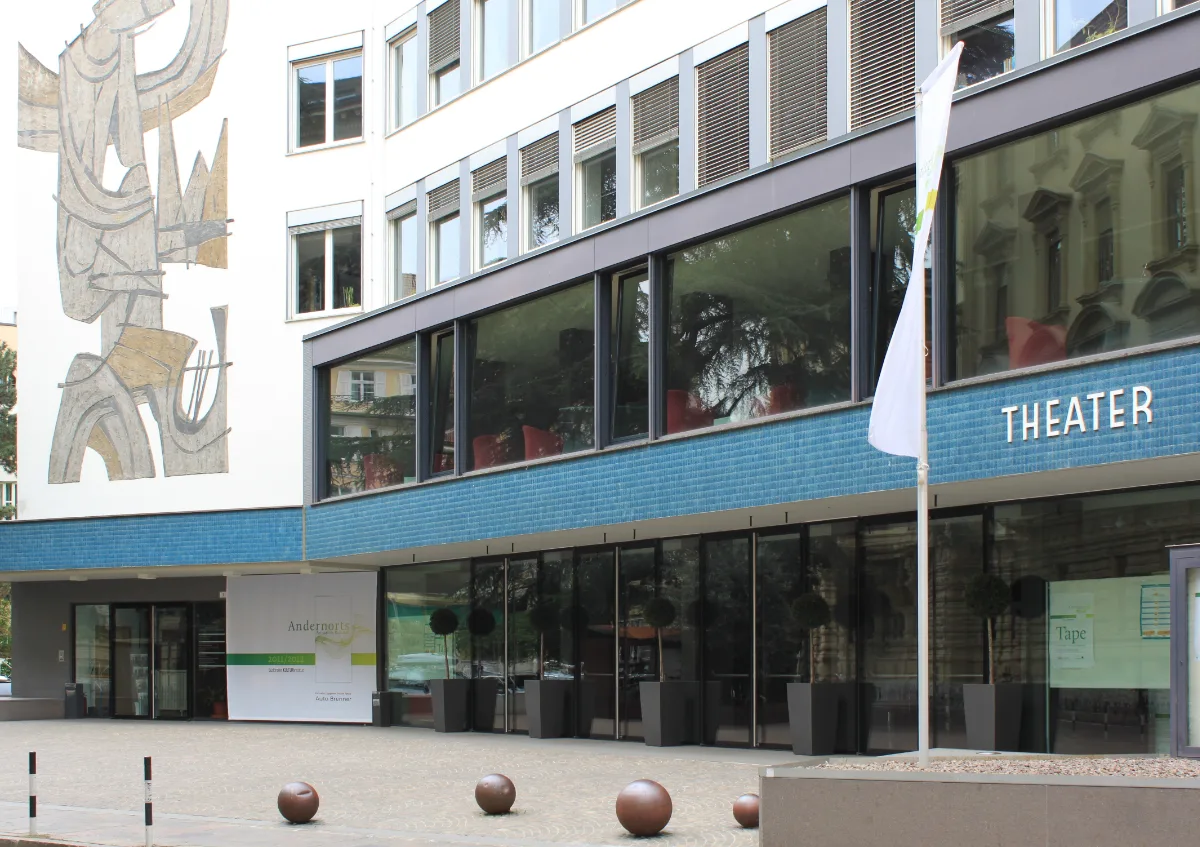 Photo des Vorplatzes eines Gebäudes. Auf dem Gebäude die Aufschrift "Theater" und ein buntes Fresko