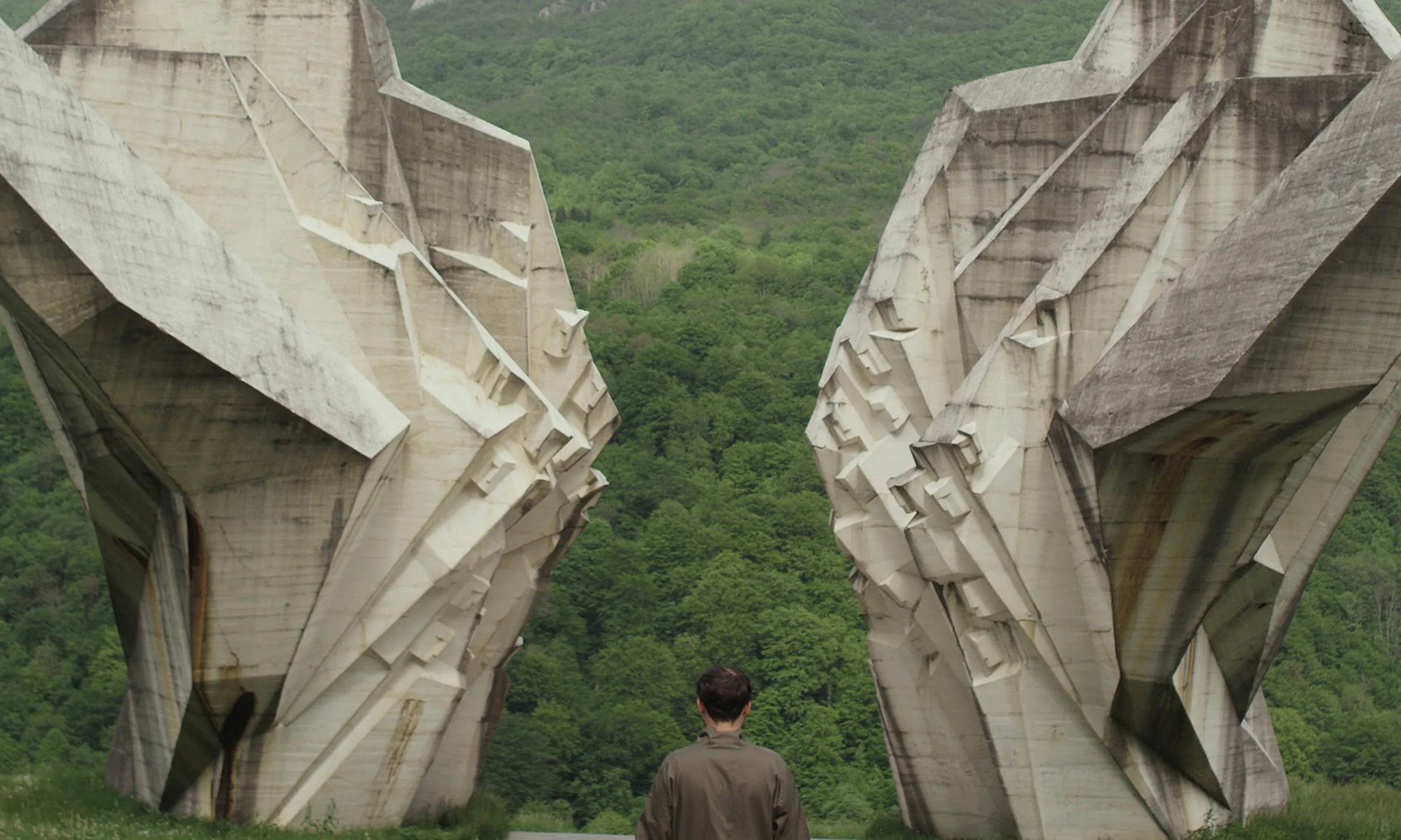 Denkmal aus Beton links und rechts, in der Mitte eine Person mit Rücken zur Kamera. Wald im Hintergrund