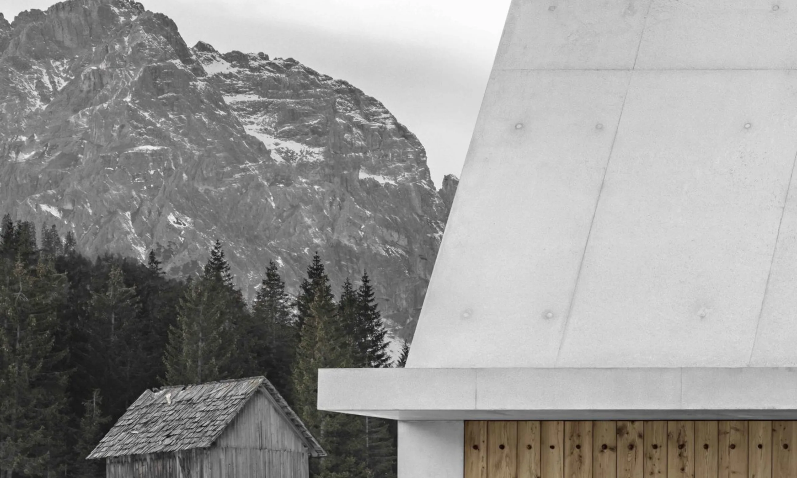 Betongebäude mit Tor aus Holz, im Hintergrund eine Scheune aus gealtertem Holz und eine alpine Landschaft