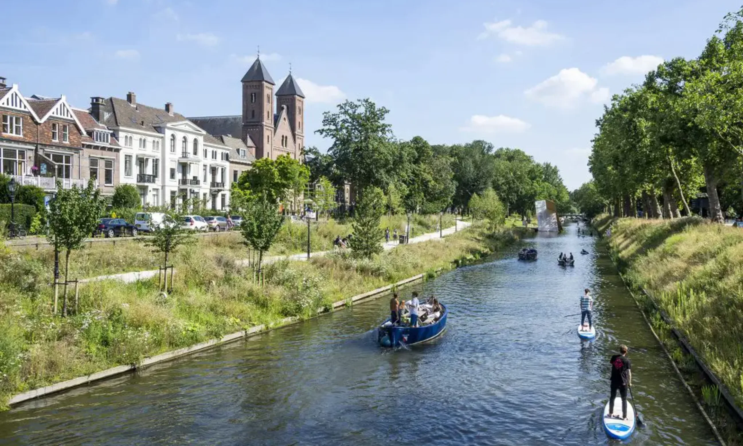 Flusslandschaft im urbanen Kontext. Menschen auf Booten, herum großzügige Grünräume