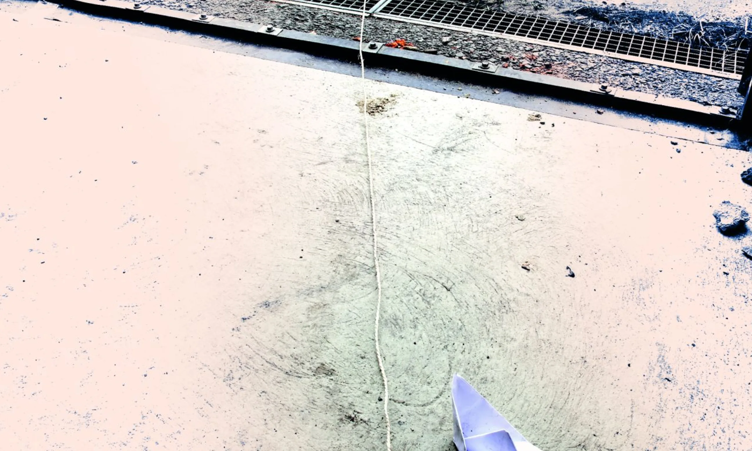 Fotografie einer Straßensituation. Ein Papierboot, eine Papierrolle verbunden durch eine Schnur mit einem Buch. In einer Pfütze spiegelt sich ein Gebäude.
