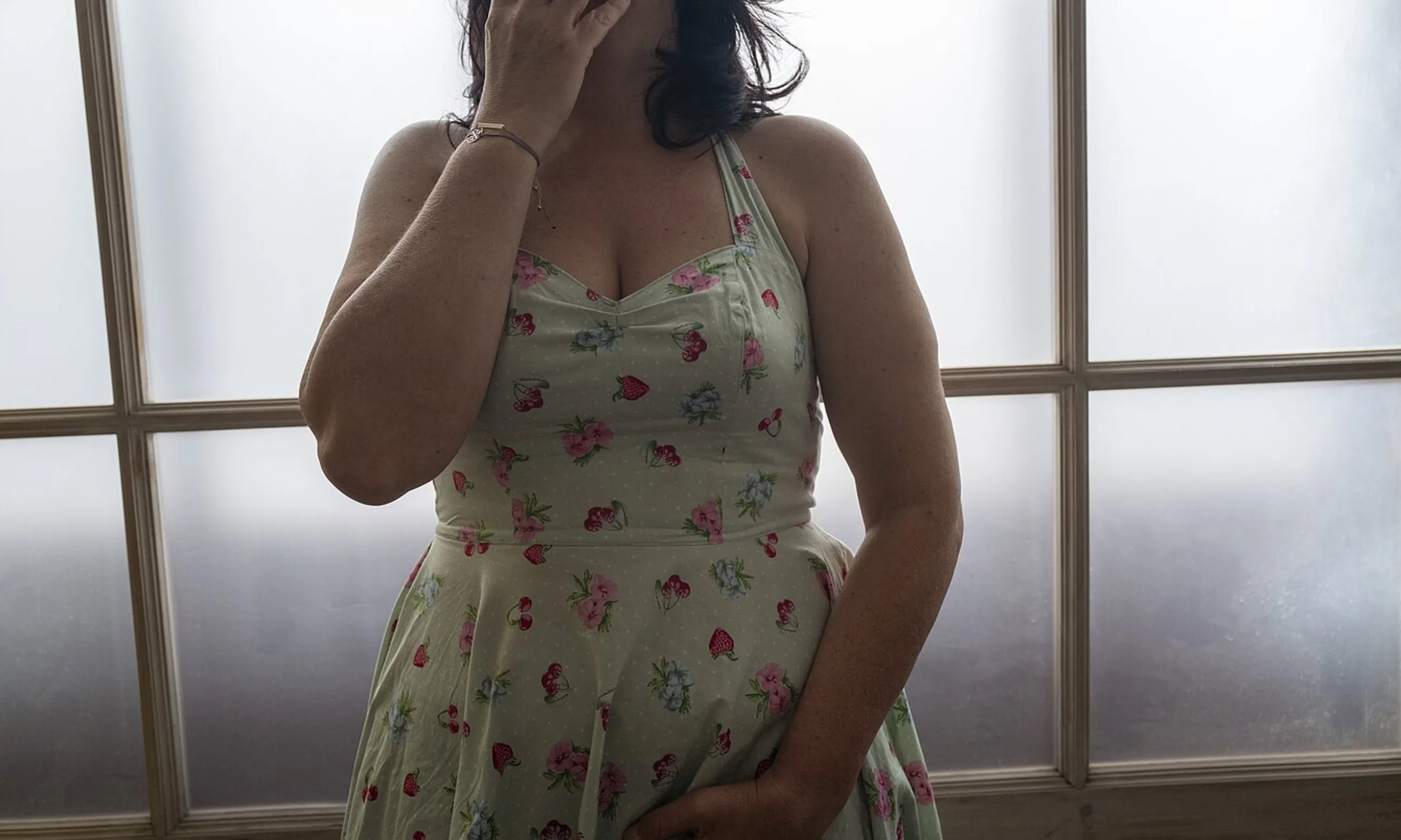 Körpermitte in einem Blumenkleid vor einer Milchglas-Wand. Die Person hält sich eine Hand im Gesicht, die Andere im Schritt
