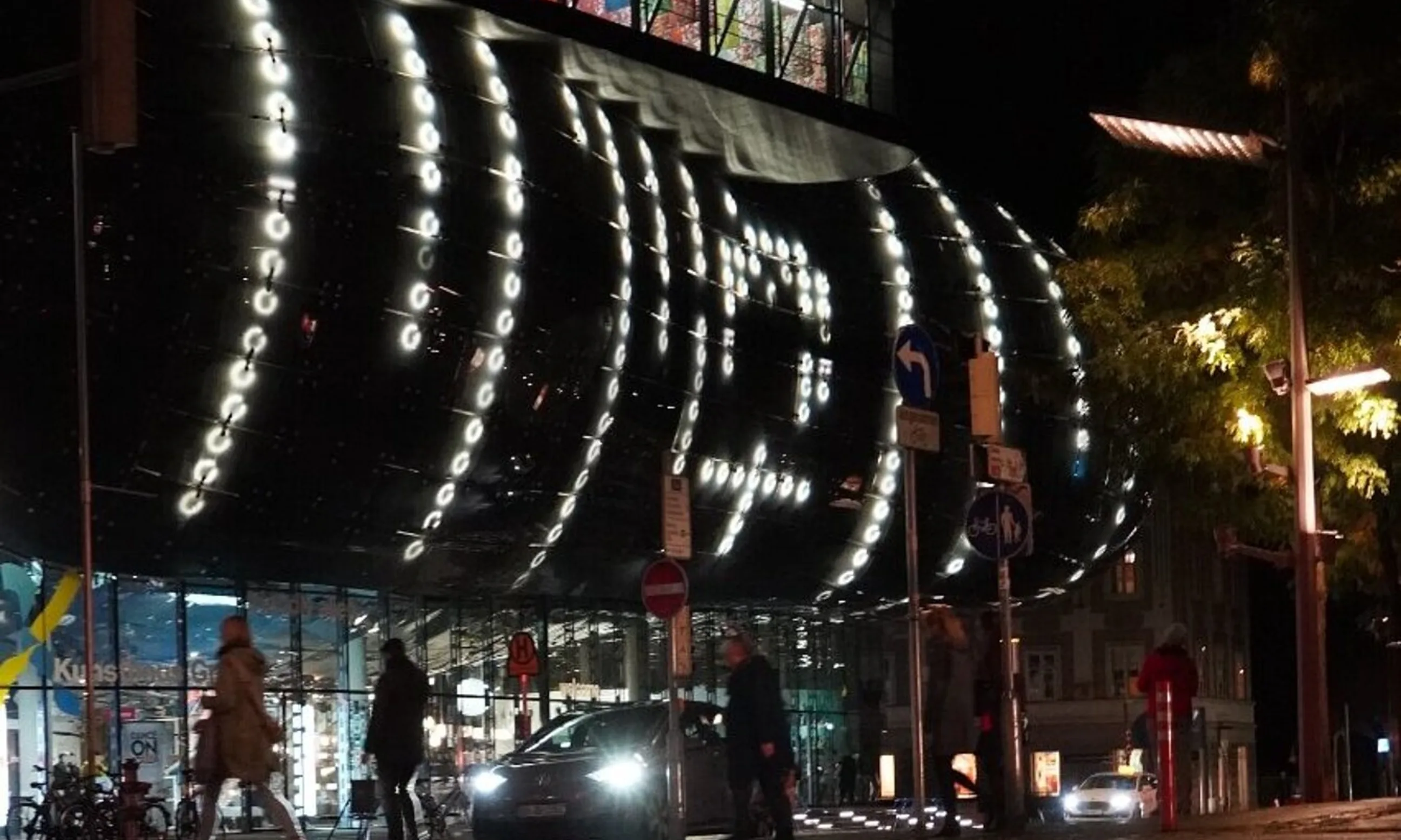 Straßenraum bei Nacht, im Hintergrund eine belichtete Glasfassade