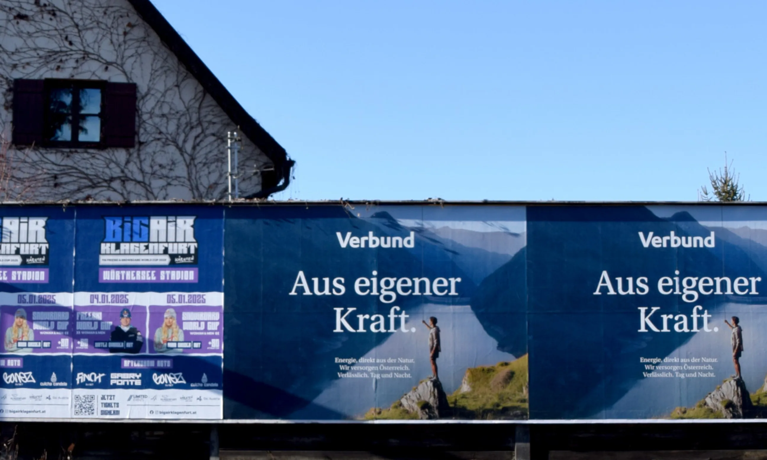Bild eines Werbepanels auf einer Lärmschutzwand, im Hintergrund der Giebel eines Hauses