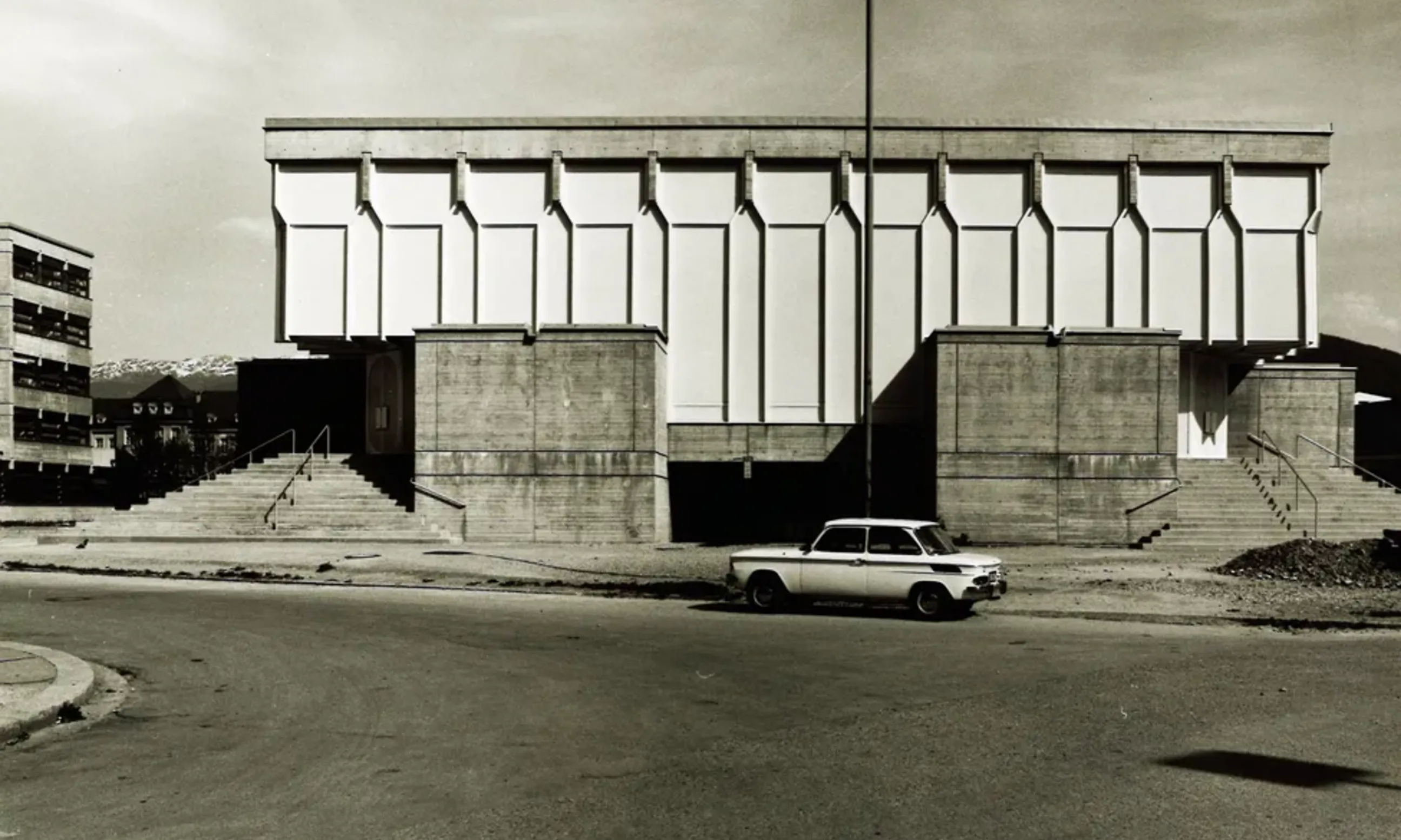 Straßenraum mit einem Auto im Vordergrun, im Hintergrund eine brutalistische Kirche