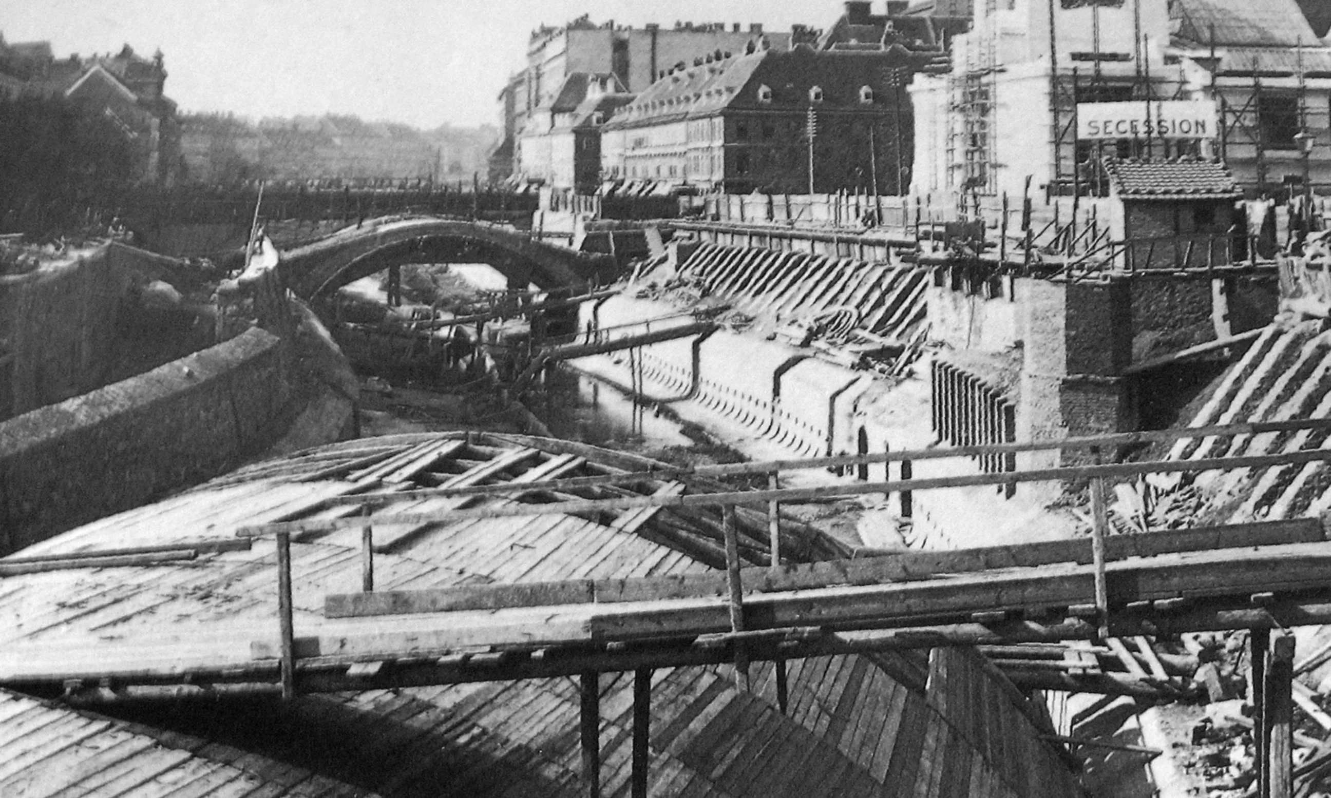 Schwarz-weißes Foto einer Baustelle, im Vordergrund ein Gewölbe, im Hintergrund mehrere Gebäude und Wasser