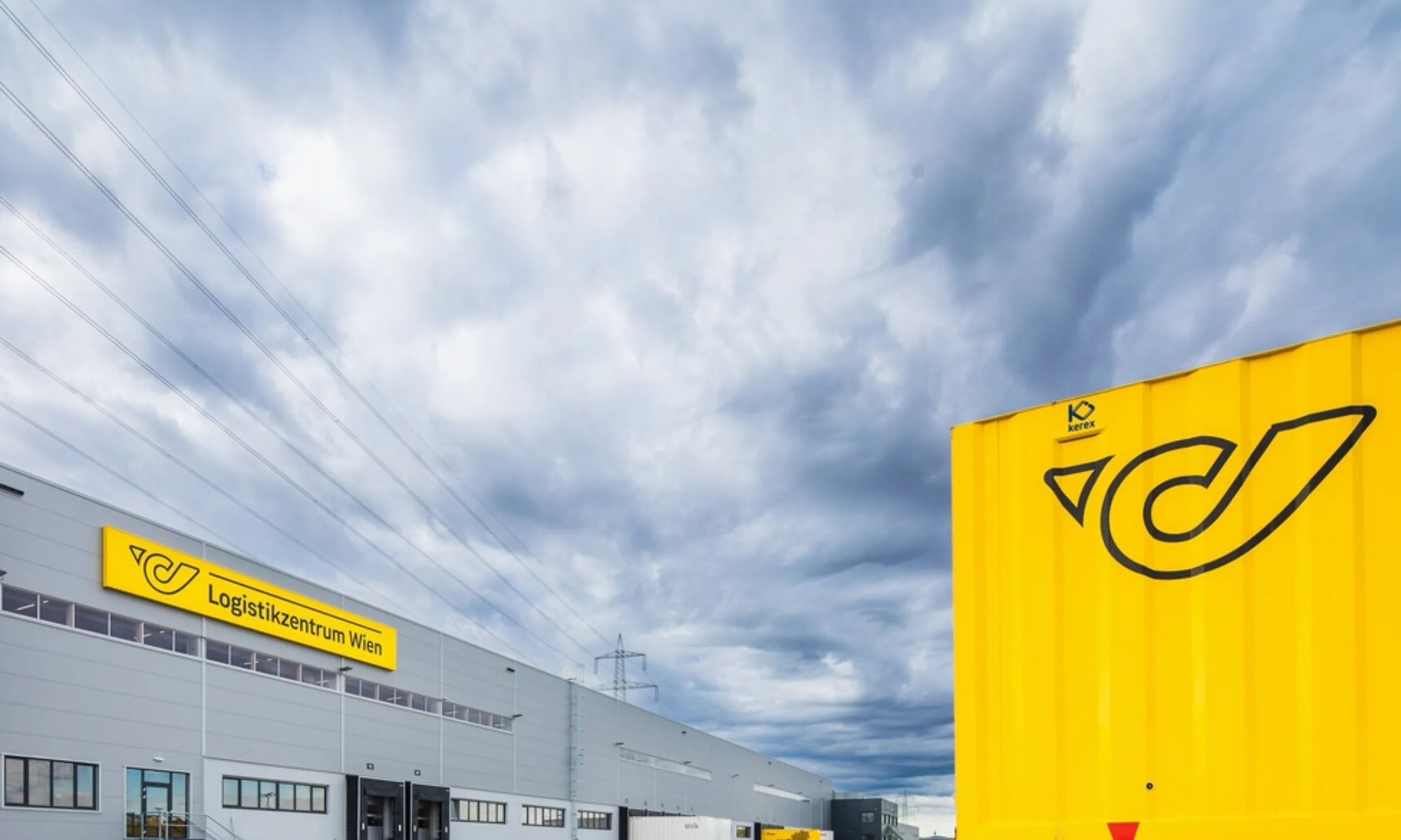 Im Vordergrund ein gelber container mit Posthorn, im Hintergrund ein graues Gebäude mit der Aufschrift Logistikzentrum Wien