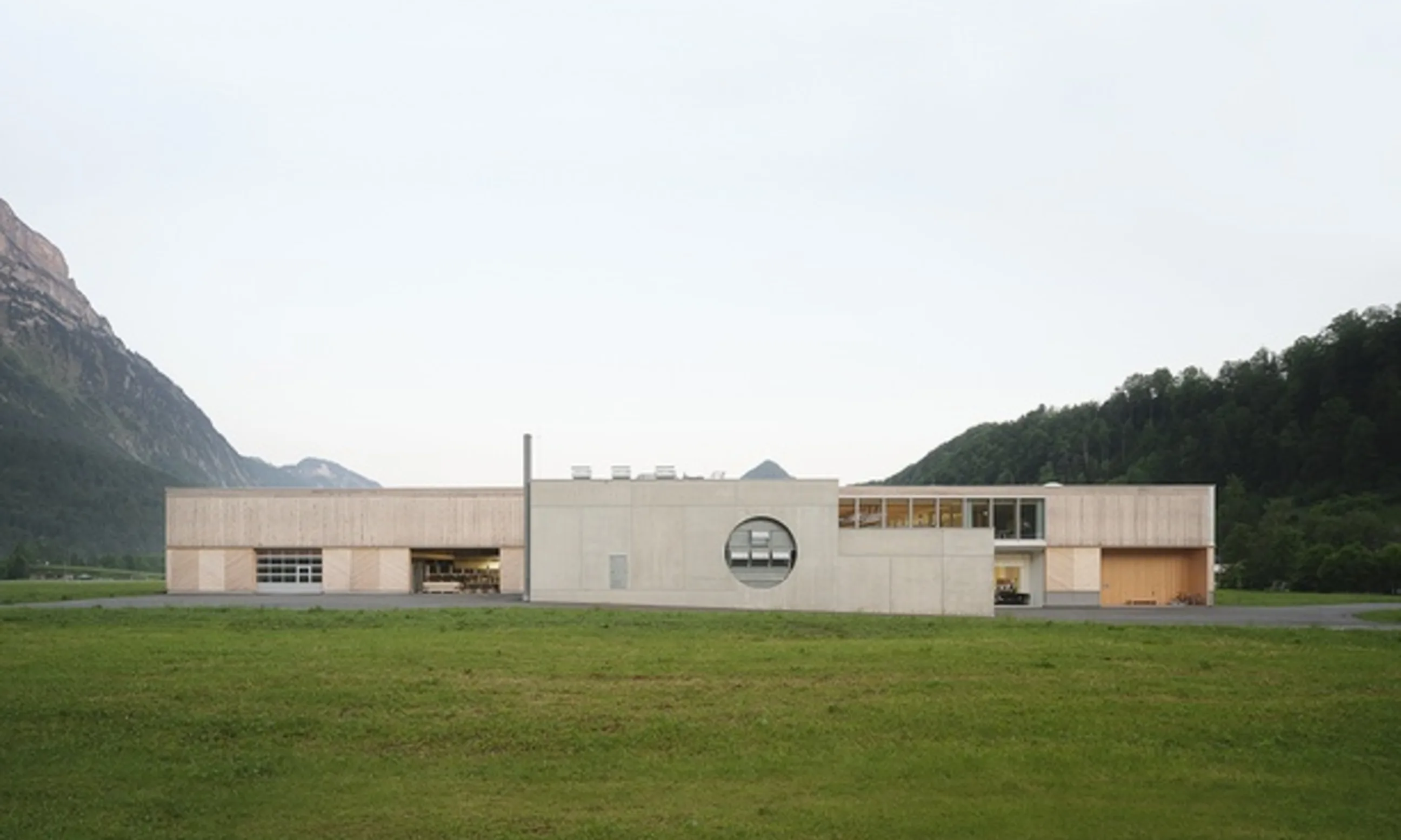 Bild einer Gebäudes aus Beton und Holz, umgeben von einer Wiese und Wäldern links und rechts