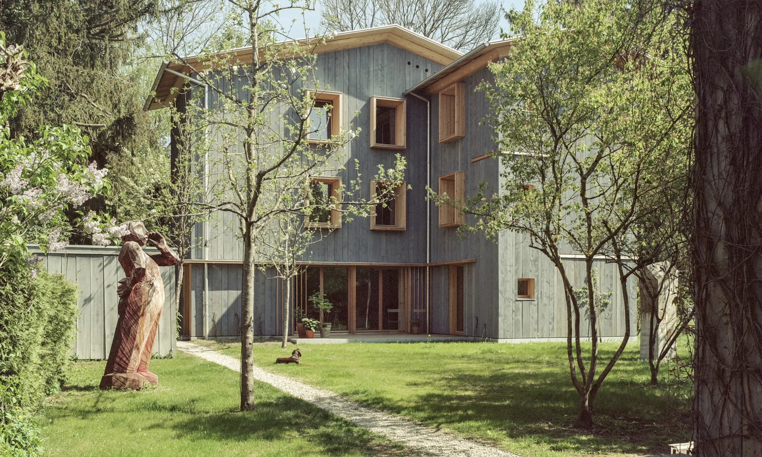Im Vordergrund eine Gartenlandschaft, im Hintergrund ein grünes Haus mit Giebeldach