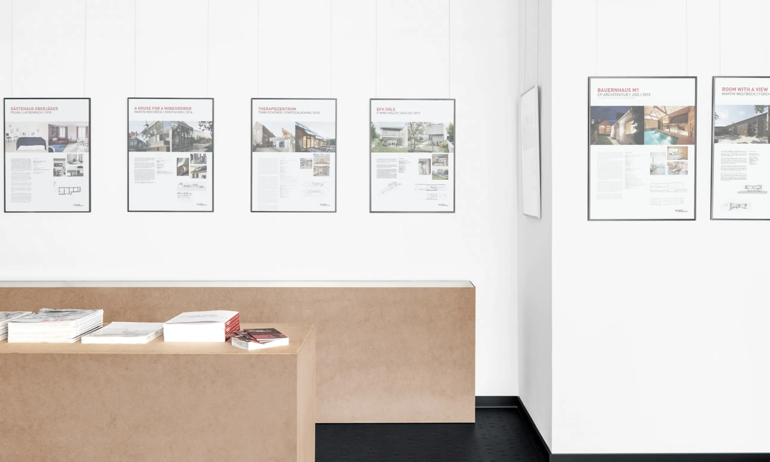 Blick in Ausstellungsraum: An der Wand hängen Bilder und Plakate von Projekten, im Raum stehen Podeste au Holz mit Büchern