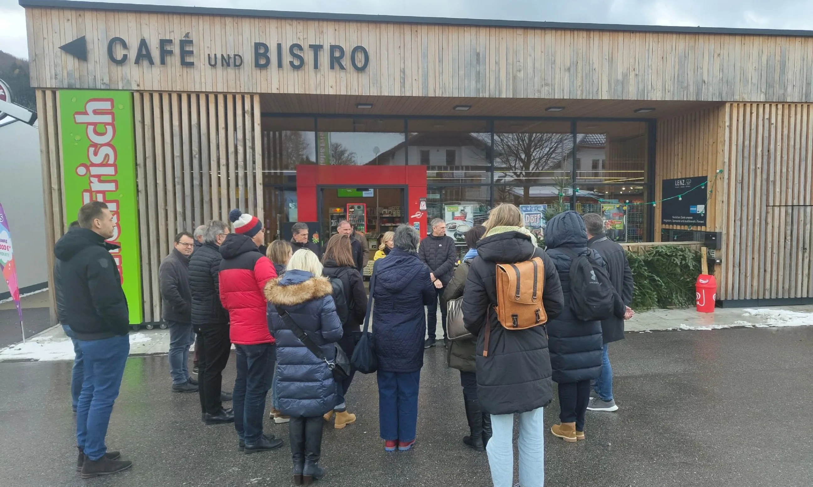 Eine Gruppe an Menschen steht vor einem eingeschossigen Gebäude mit Holzfassade