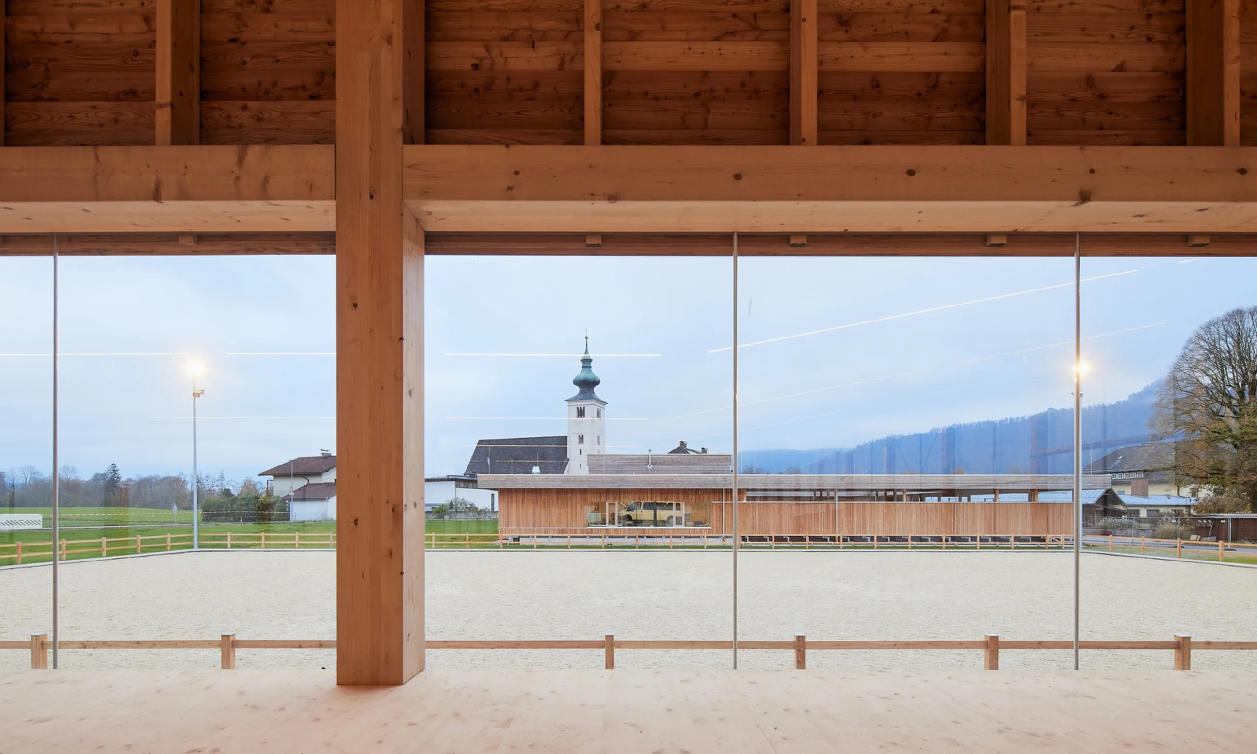 Im Vordergrund eine Halle aus Holz, durch die Glasfassade im Hintergrund ist eine Dorflandschaft sichtbar
