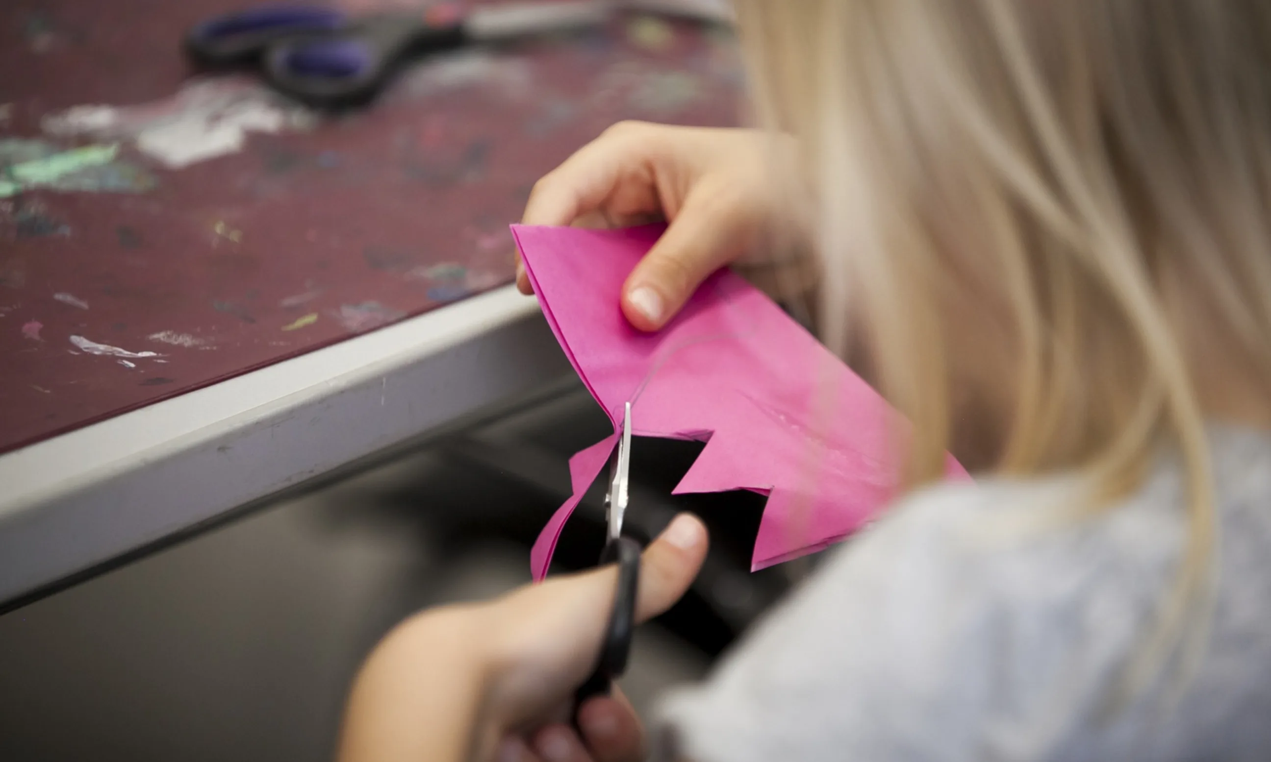 Photo eines Kindes, welches dabei ist, mit einer Schere ein pinkes Stück Papier zu schneiden