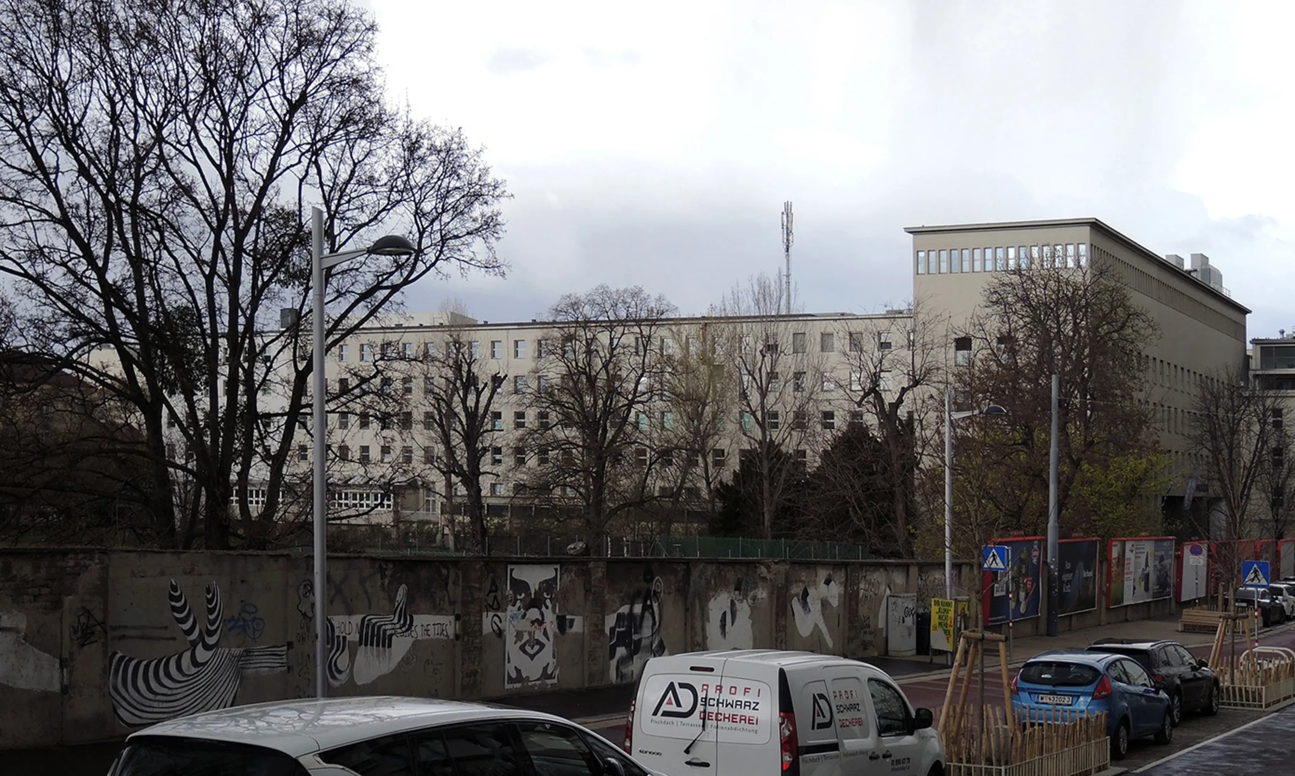 Bild einer Straße mit geparkten autos, im Hitnergrund ein weißes mehrgeschossiges Gebäude