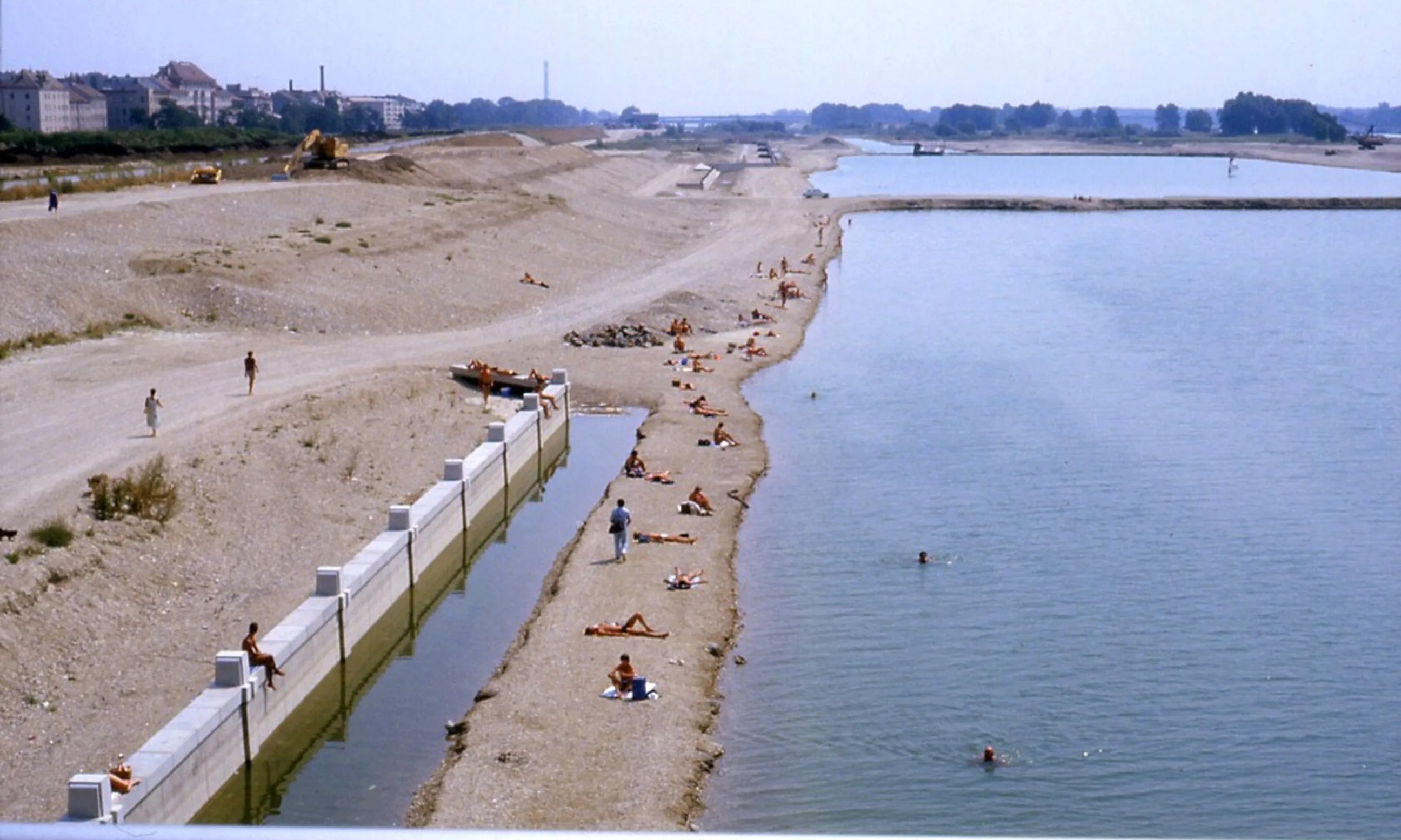 Bild eines Flusstrandes, Menschen liegen in der Sonne oder schwimmen im Fluss. Im Hintergrund die Silhouette eines bebauten Gebiets
