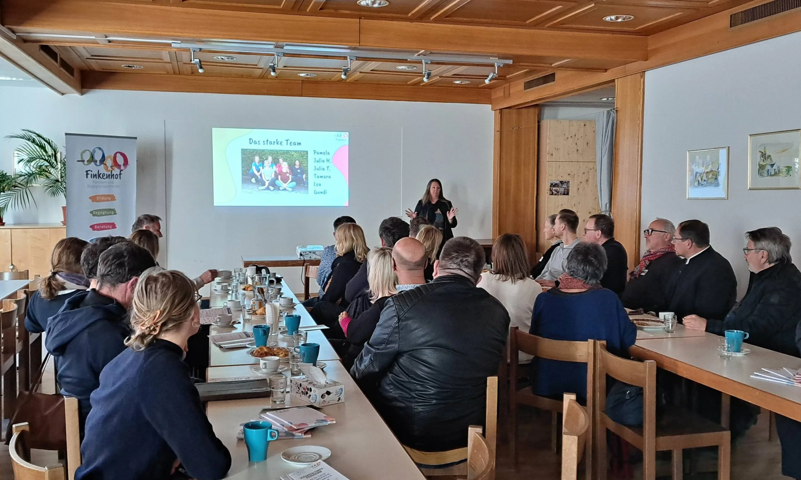 Aufnahme eines Vortrags in einem Innenraum mit vielen Menschen im Publikum und einer Vortragenden im Hintergrund. Hinter ihr eine Projektionswand