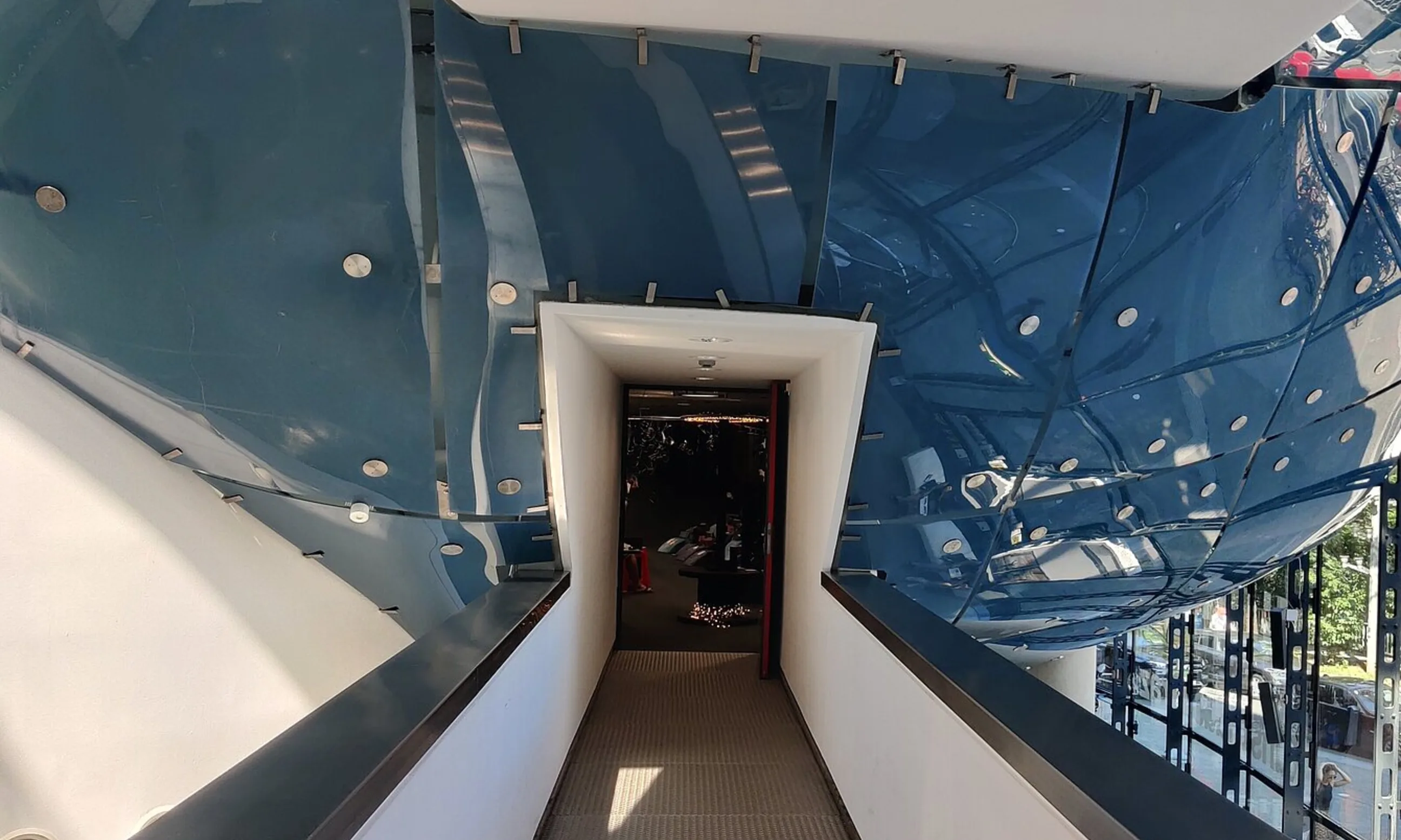Eine weiße Brücke führt in eine blaue Glaswand, in der Mitte eine offene Tür