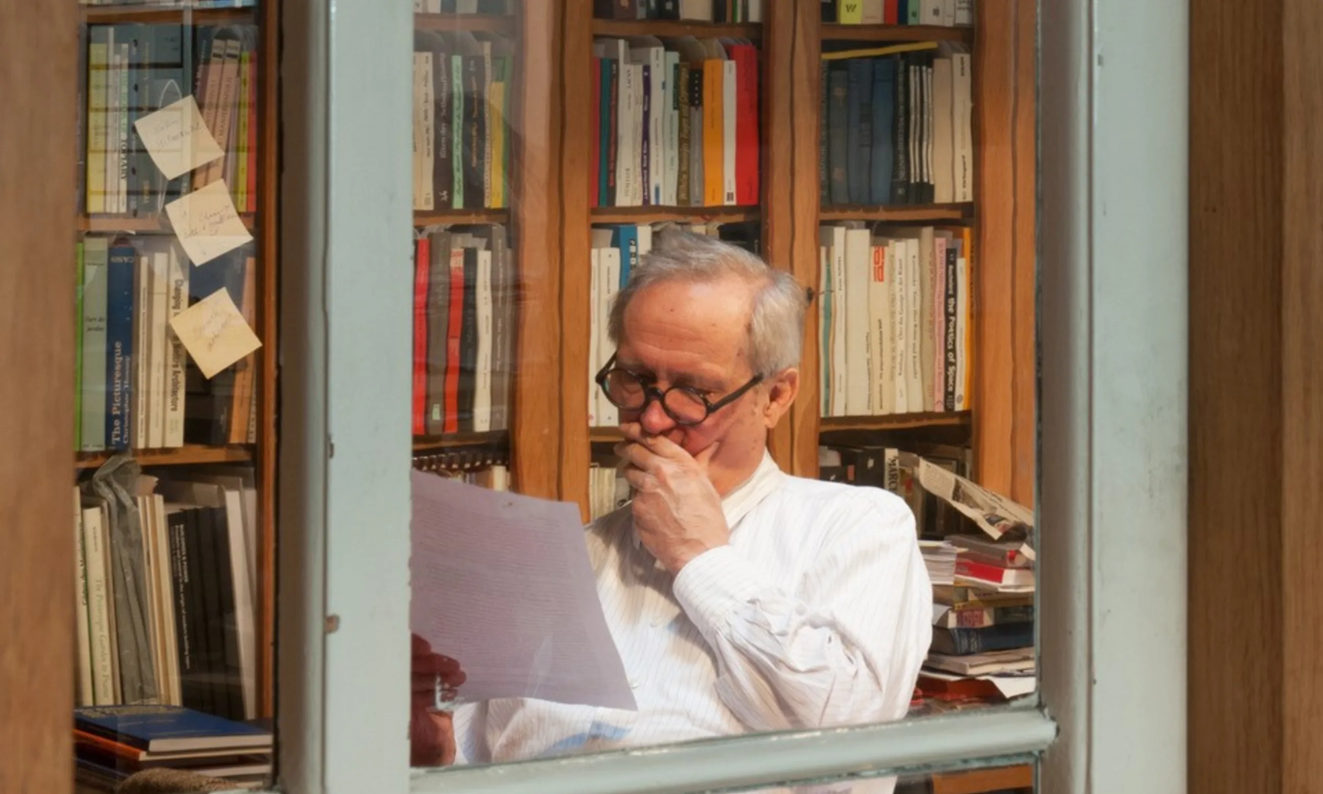 Porträt eines Mannes an seinem Schreibtisch, umgeben von Büchern und Bücherregalen, fotografiert durch ein Fenster