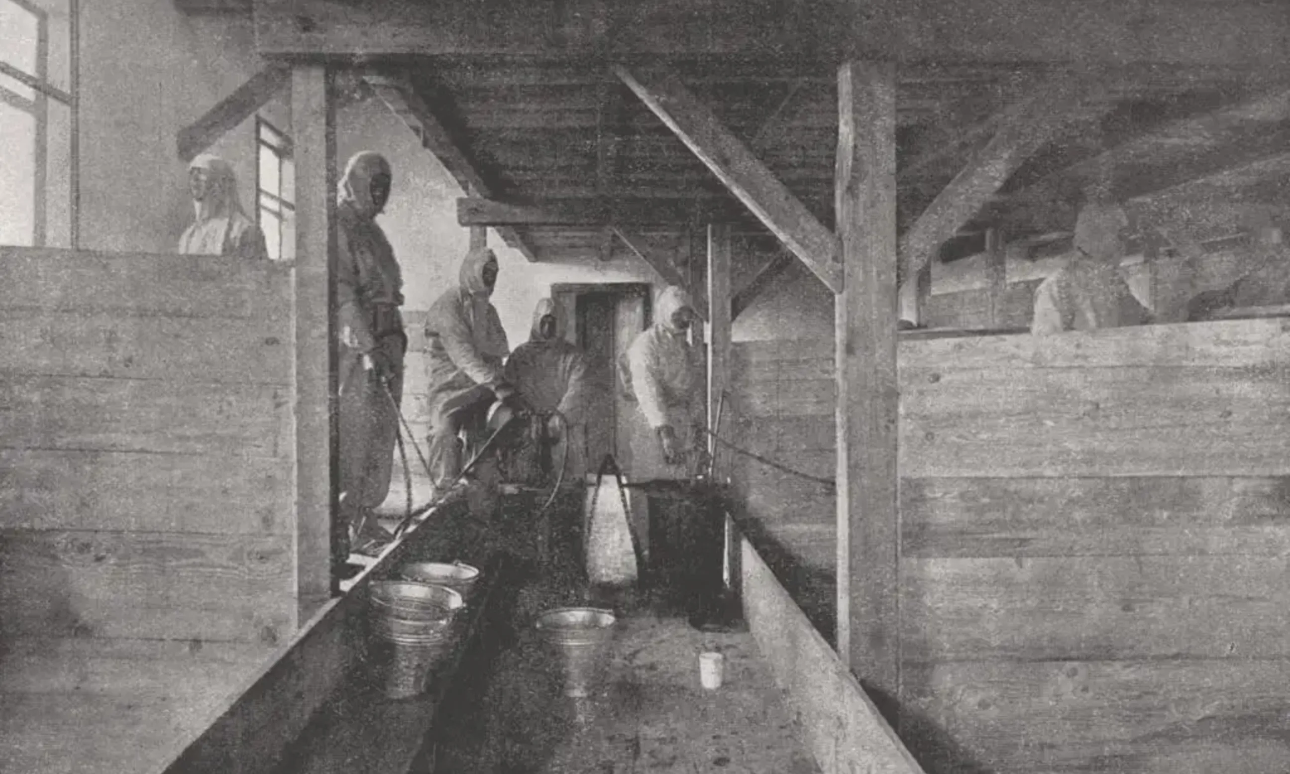 Schwarz-weißes Foto des Inneren eines Holzgebäudes, im vordergrund 5 Menschen in Schutzausrüstung, bei Reinigungsarbeiten dokumentiert