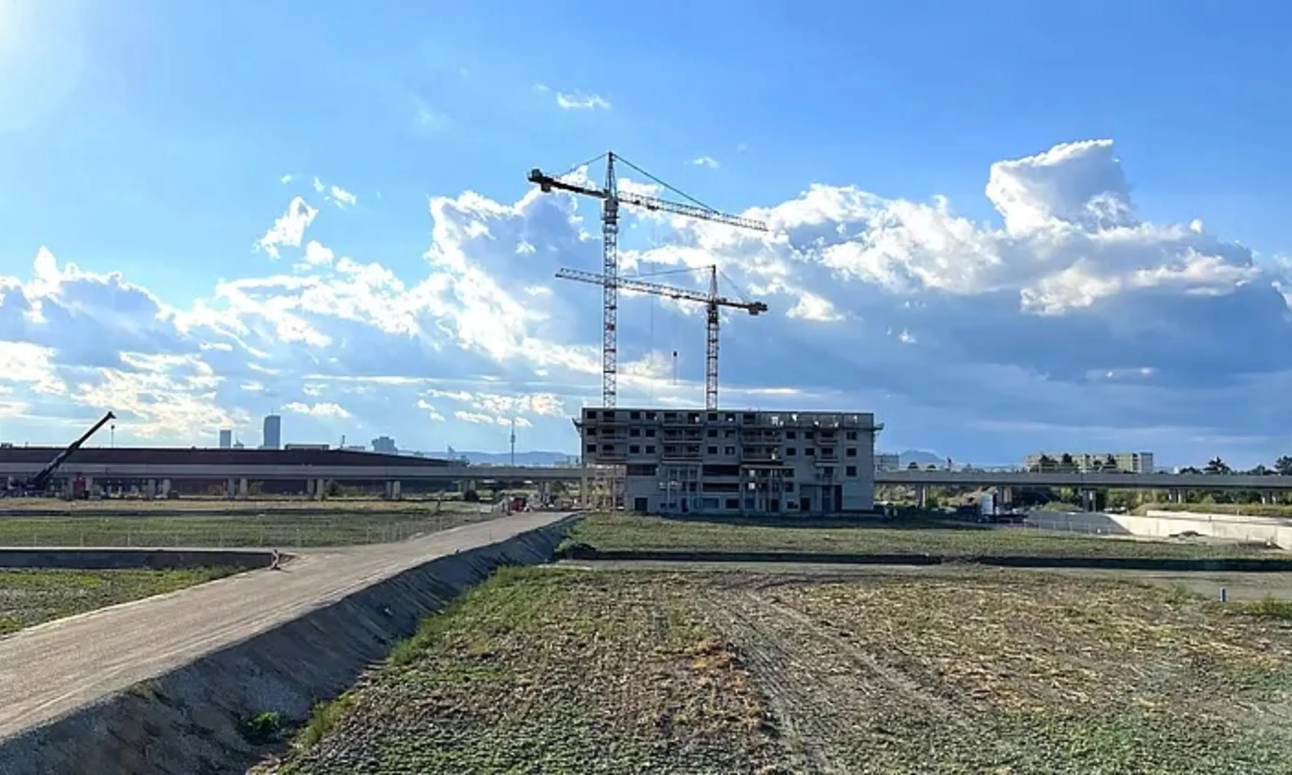 Eine Wiese mit links seitlich einer unasphaltierten Straße, die zu einem Rohbau führt