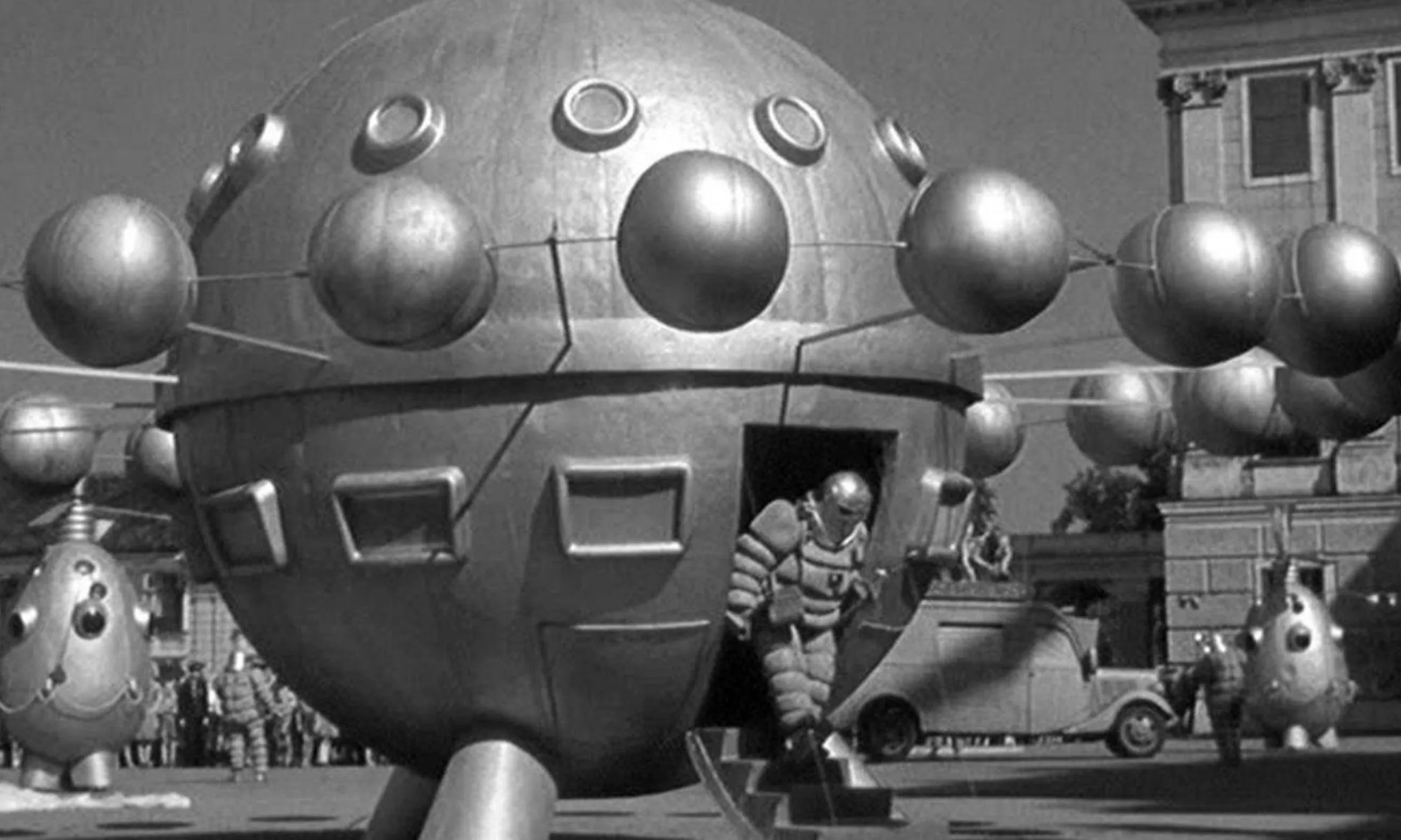 Filmstill in schwarz-weiß, Mensch steigt im Weltraumanzug aus silberner Kugel aus, im Hintergrund die historische Kulisse einer Stadt