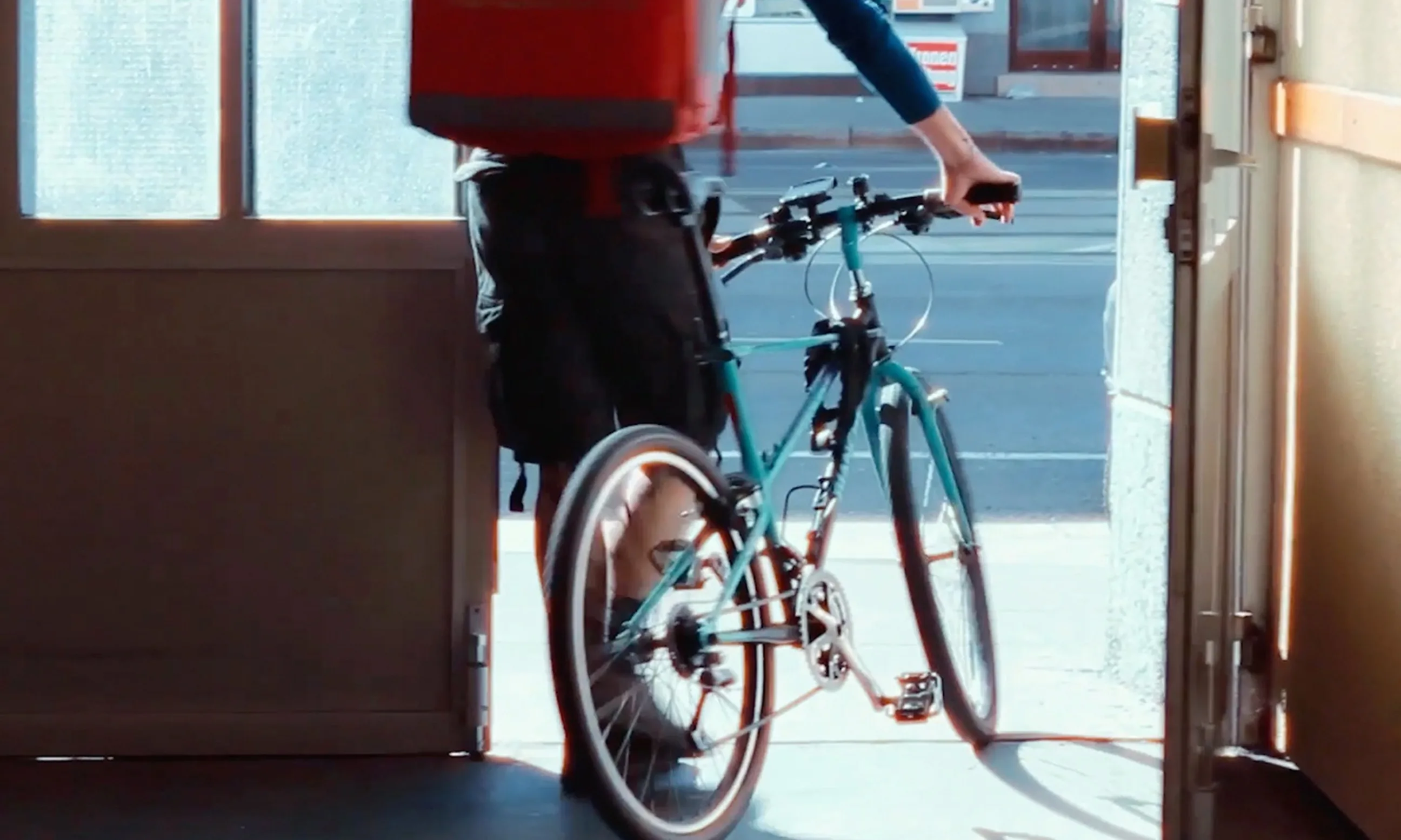 Ein Mensch mit Rucksack und Rad spaziert durch ein Tor auf die Straße, Ansicht von hinten