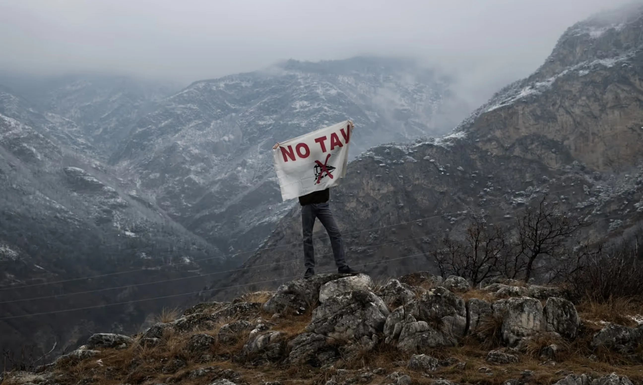 Bild einer Person in einer nebligen alpinen Landschaft, die mit dem Rücken zur Kamera steht und eine Flagge mit der Aufschrift NO TAV in die Kamera hält