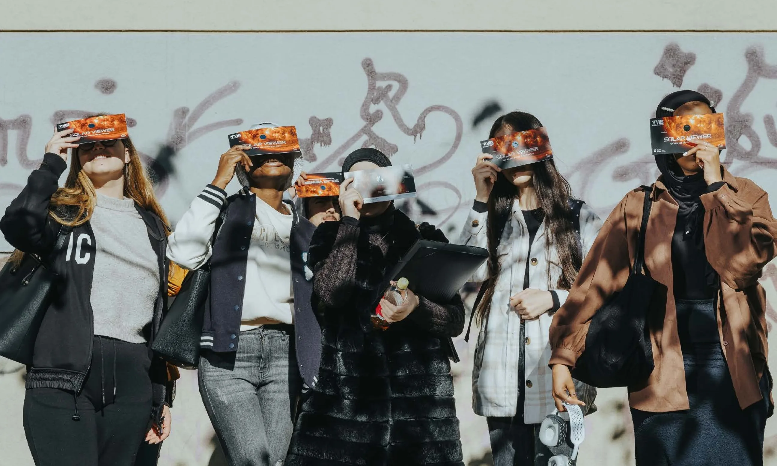 Eine Gruppe von fünf Personen steht an einer Wand und blickt nach oben, sie halten ihre Gesichter bedeckt