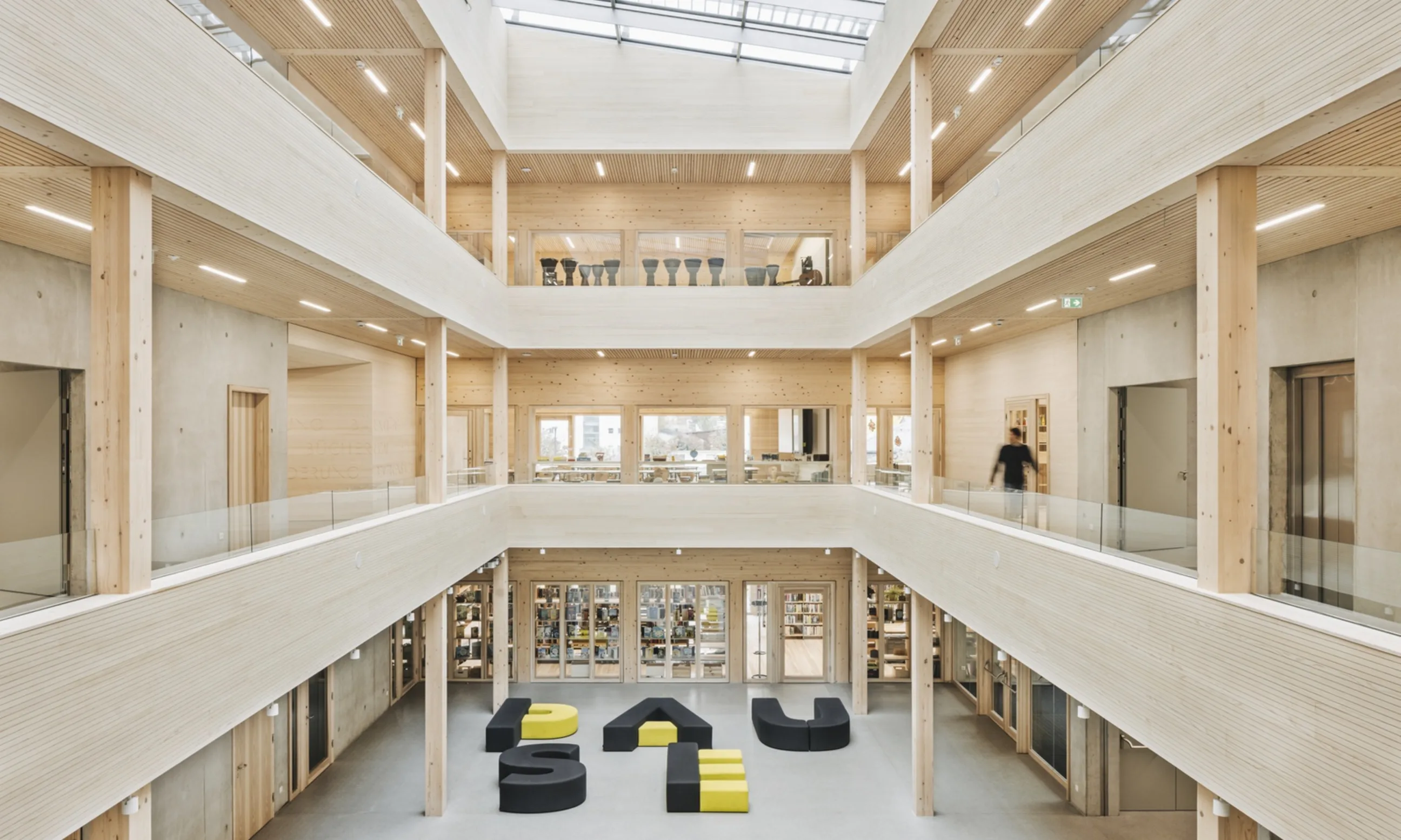 Photo eines Atriums eines mehrgeschossigen Gebäudes. Mittig im Erdgeschoß eine Sitzlandschaft dessen Elemente das Wort "Pause" ergeben