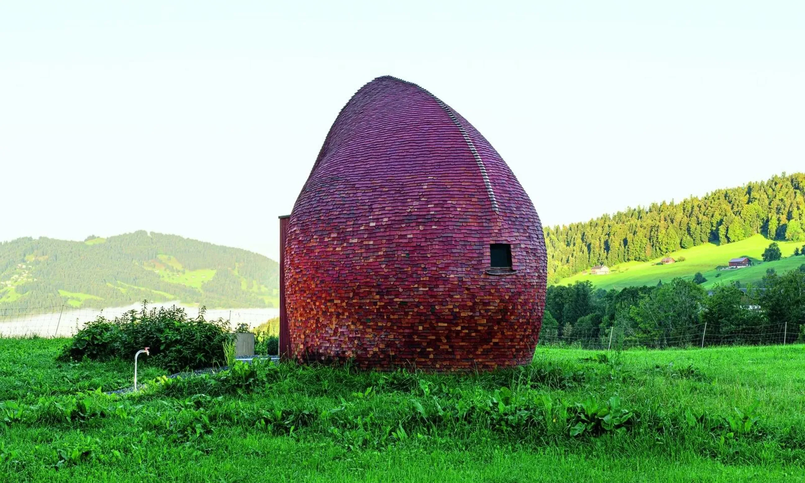 Ein rotes, rundes Gebäude steht auf einer grünen Wiese, im Hintergrund eine hügelige Landschaft
