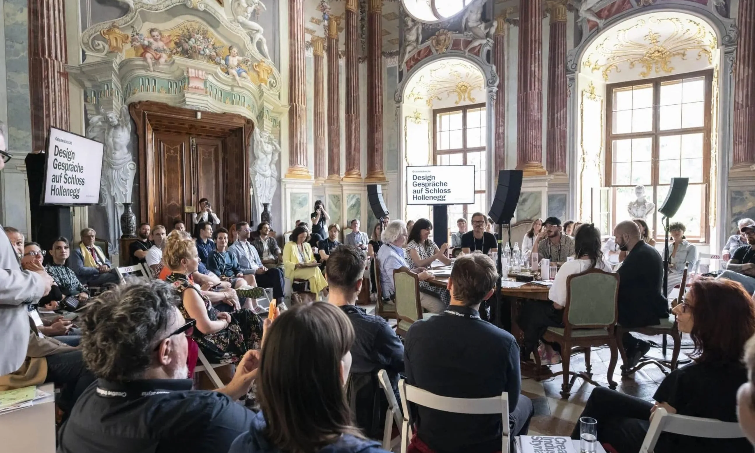 Ein Raum voll mit sitzenden Menschen, im Mittelpunkt ein runder Tisch