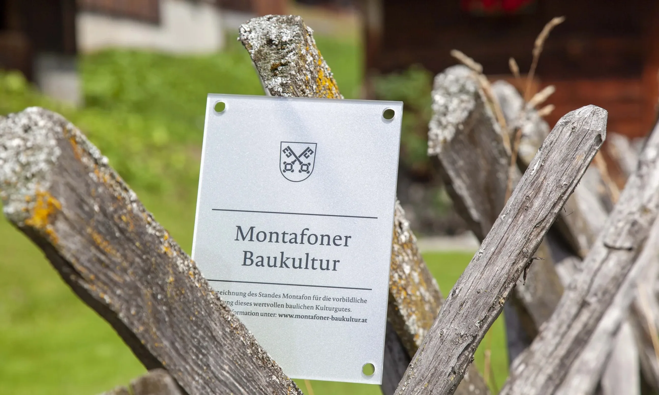 Foto einer Plakette mit der Aufschrift Montafoner Baukultur, an einen hölzernen Zaun angelehnt